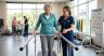 A photorealistic composite scene of a modern rehabilitation clinic, set against a background view of a park and pond through a large window. In the center, a friendly male therapist in a blue polo shirt (labeled Ascend Rehabilitation) stands beside an older woman in a grey shirt, watching her as she carefully cuts red bell peppers on a wooden cutting board with a large knife, practicing a practical daily life task. On a nearby desk, a progress chart with an upward trend is visible. In the background, another therapist and a senior man are interacting at a table (likely speech therapy), with a 'Progress Chart' and wall plaque for 'ASCEND REHABILITATION - (Fictional)' on the wall. A bottom-right inset image shows essential recovery paperwork: a 'MEDICARE Part B' card, a clipboard with a 'SUPPLEMENTAL INSURANCE FORM', and other insurance documents, linking the clinic's physical activity to the coverage options discussed in the preceding infographic.